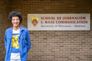 RadioLab co-host Latif Nasser stands outside Vilas Hall on the UW–Madison campus. He smiles in the foreground; in the background, there is a white sign reading, "School of Journalism & Mass Communication, University of Wisconsin–Madison."