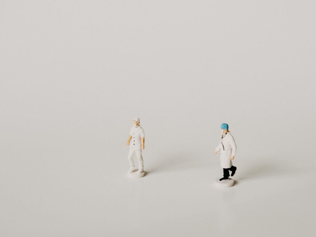 Photo of two plastic figurine doctors positioned to look as if they are walking in the same direction, on a stark white background. 
