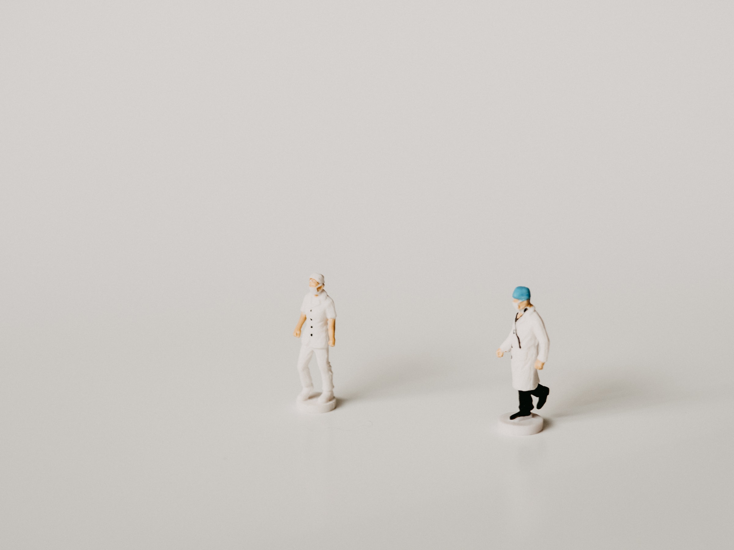 Photo of two plastic figurine doctors (white, male), posed as if to look like they are walking in the same direction, against a stark white background.