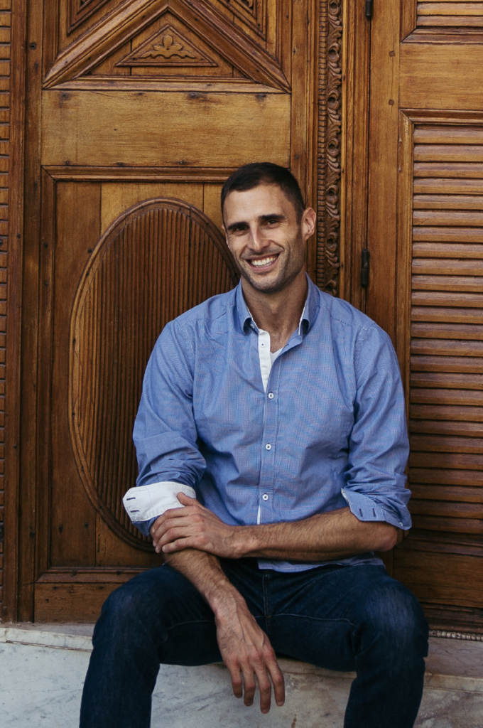 Photo of Jacob Kushner earring blue button down shirt and jeans, seated in front of large wooden doors.