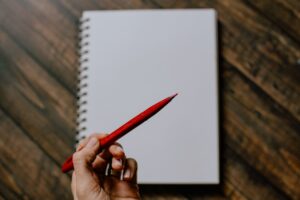 Image of a notebook opened to a blank page with a hand holding a red pencil over it.