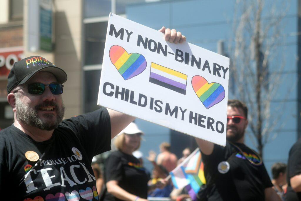 A white man with a beard in a "Pride" hat and sunglasses holds up a hand-written sign reading, "My non-binary child is my hero." The sign includes two rainbow-striped hearts and a non-binary pride flag with yellow, white, purple and black stripes. 