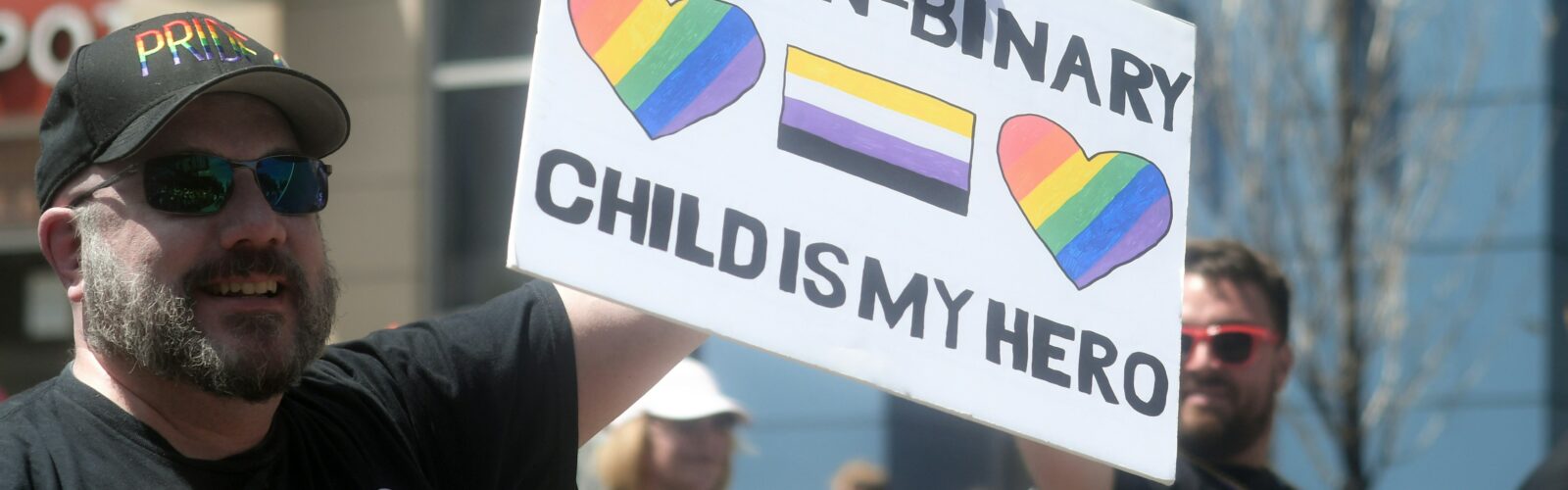 A white man with a beard in a "Pride" hat and sunglasses holds up a hand-written sign reading, "My non-binary child is my hero." The sign includes two rainbow-striped hearts and a non-binary pride flag with yellow, white, purple and black stripes.